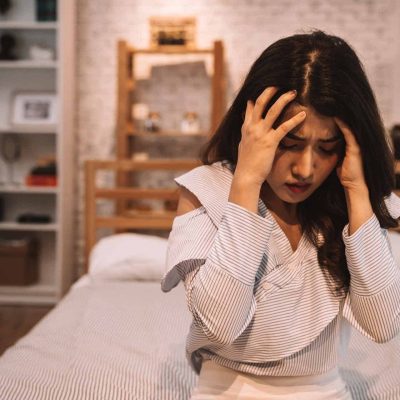 Young beautiful Asian woman having a headache while lying on bed, feeling sad and stressed at home. Disappointed and desperate girl crying.